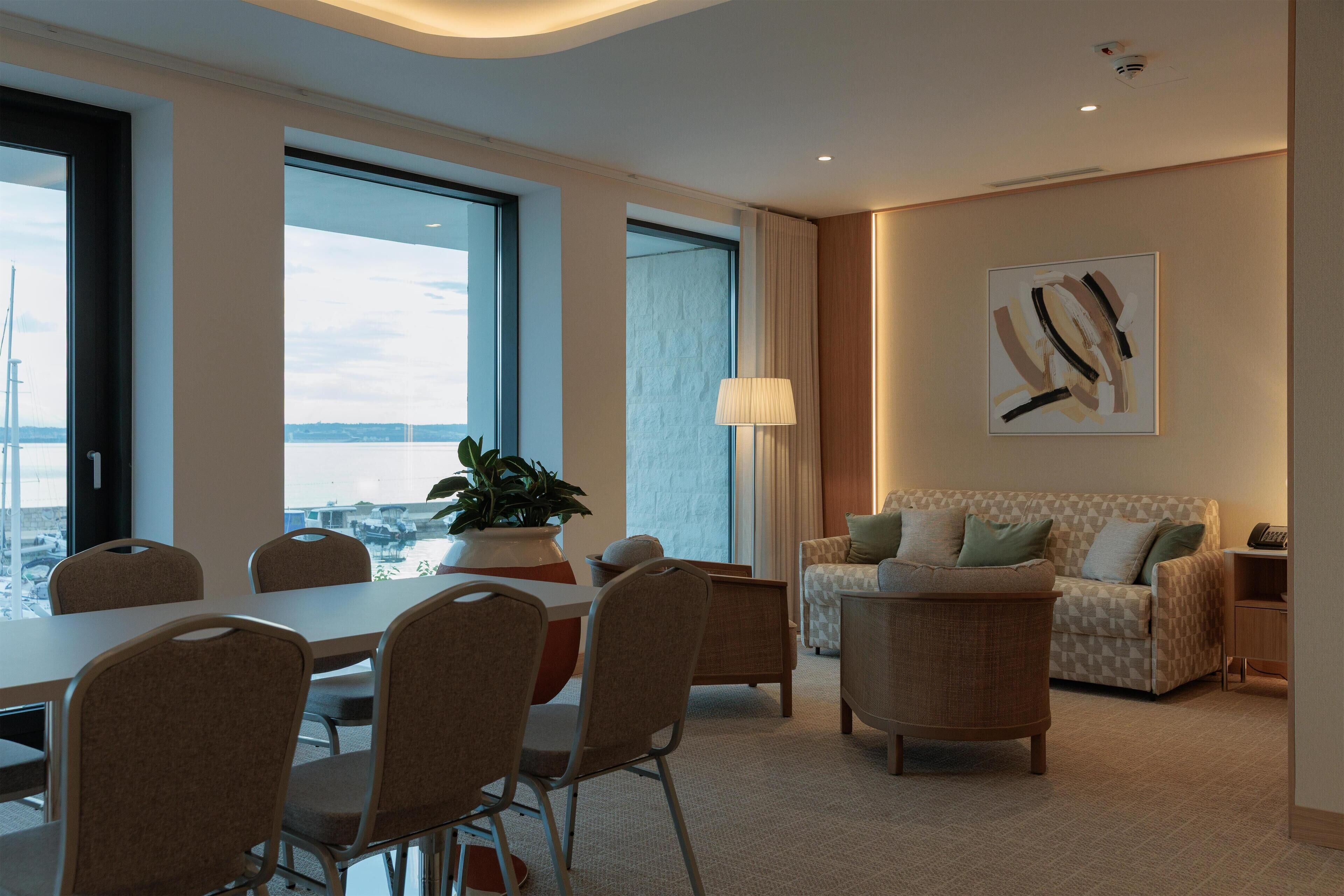 A modern hotel room with a dining area, a sofa set, and a large window offering a city view.