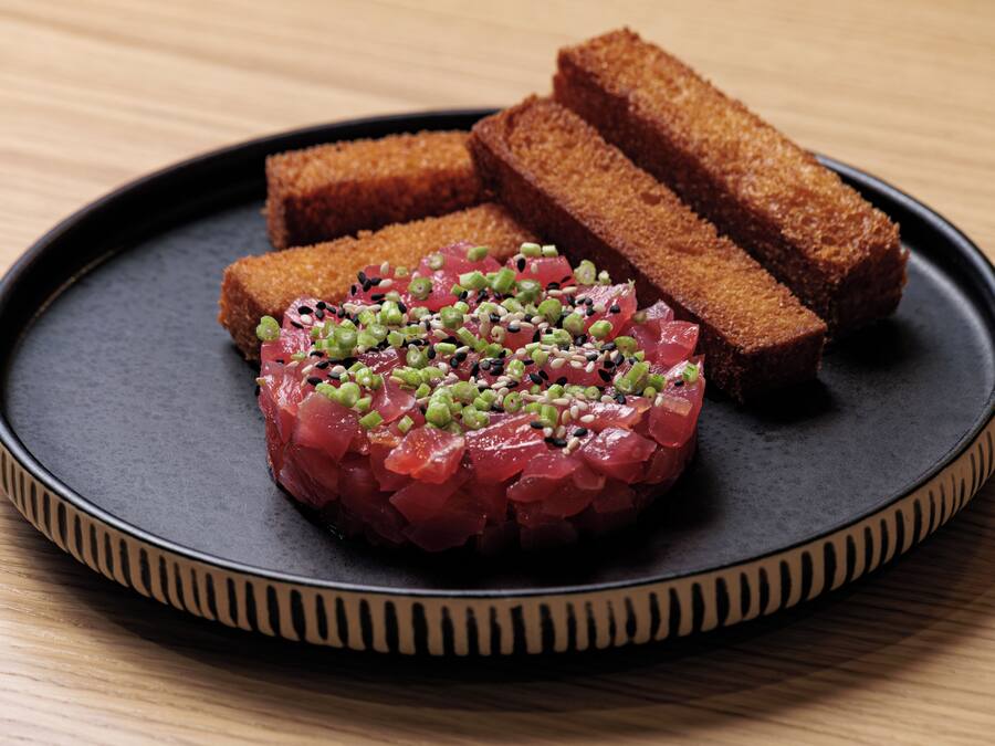 A plate with a dish of raw fish topped with green seeds and sesame seeds, accompanied by two pieces of bread on a black plate.
