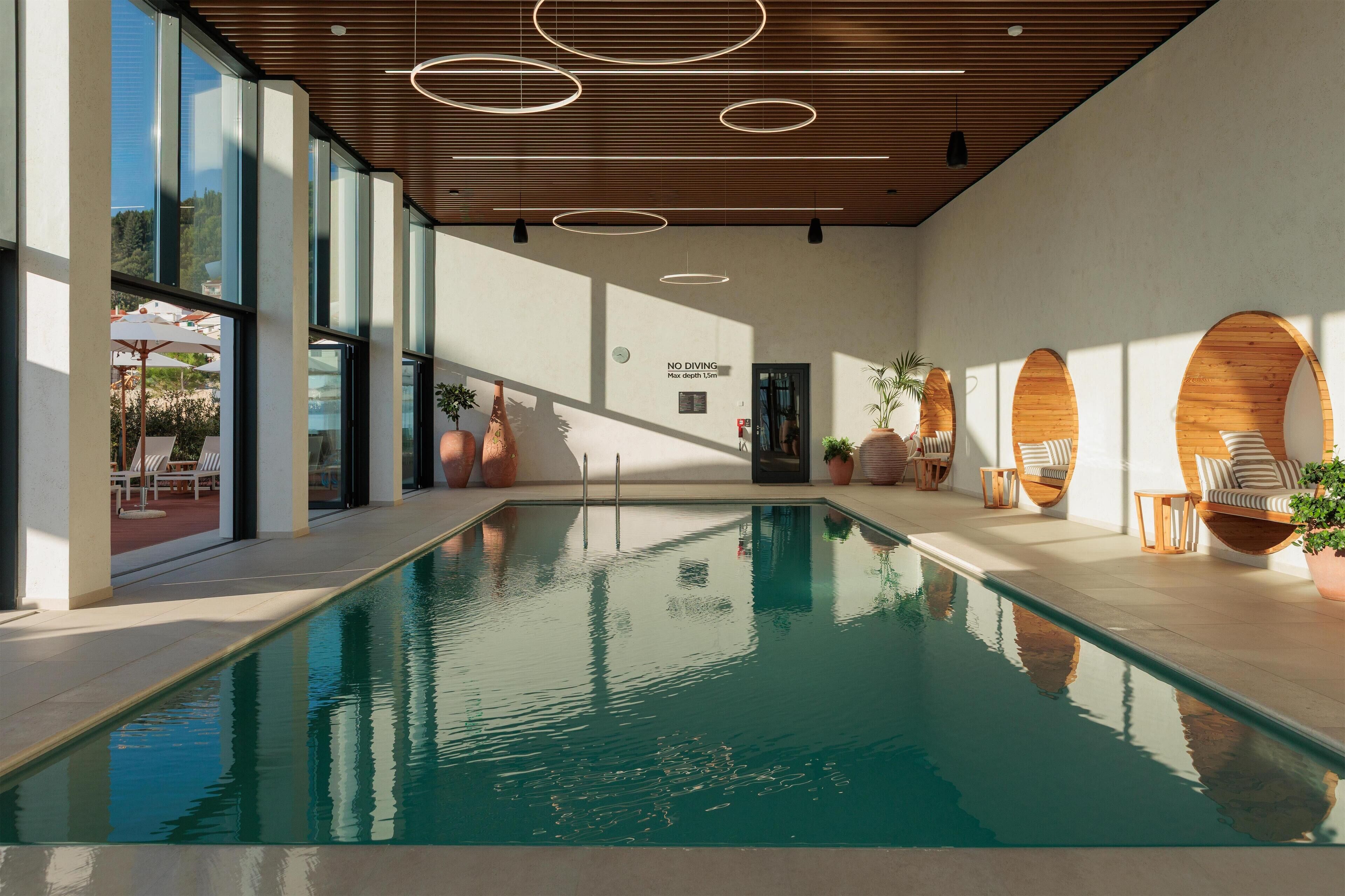 A modern indoor pool area with clear glass walls, wooden ceiling, and sunlit seating areas.
