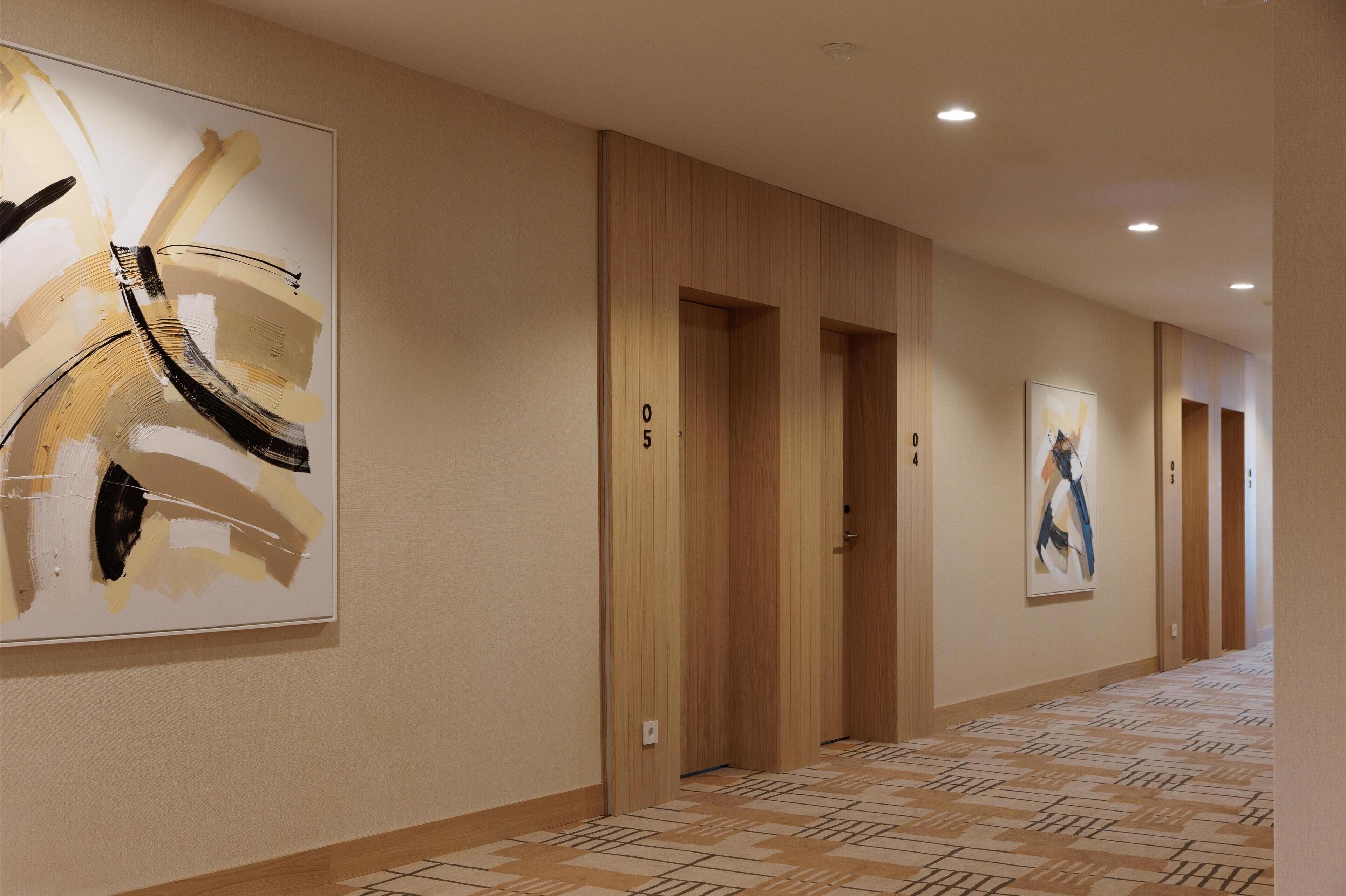 A hotel hallway with wooden doors numbered 0 to 5, a patterned carpet, and abstract art on the walls.