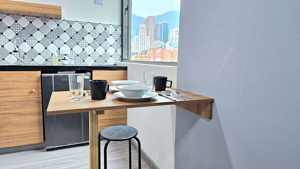 A compact kitchen with a wooden countertop, a black chair, and a window with a cityscape view. - Hotel Ktaleya Apartamentos (Bogotá)