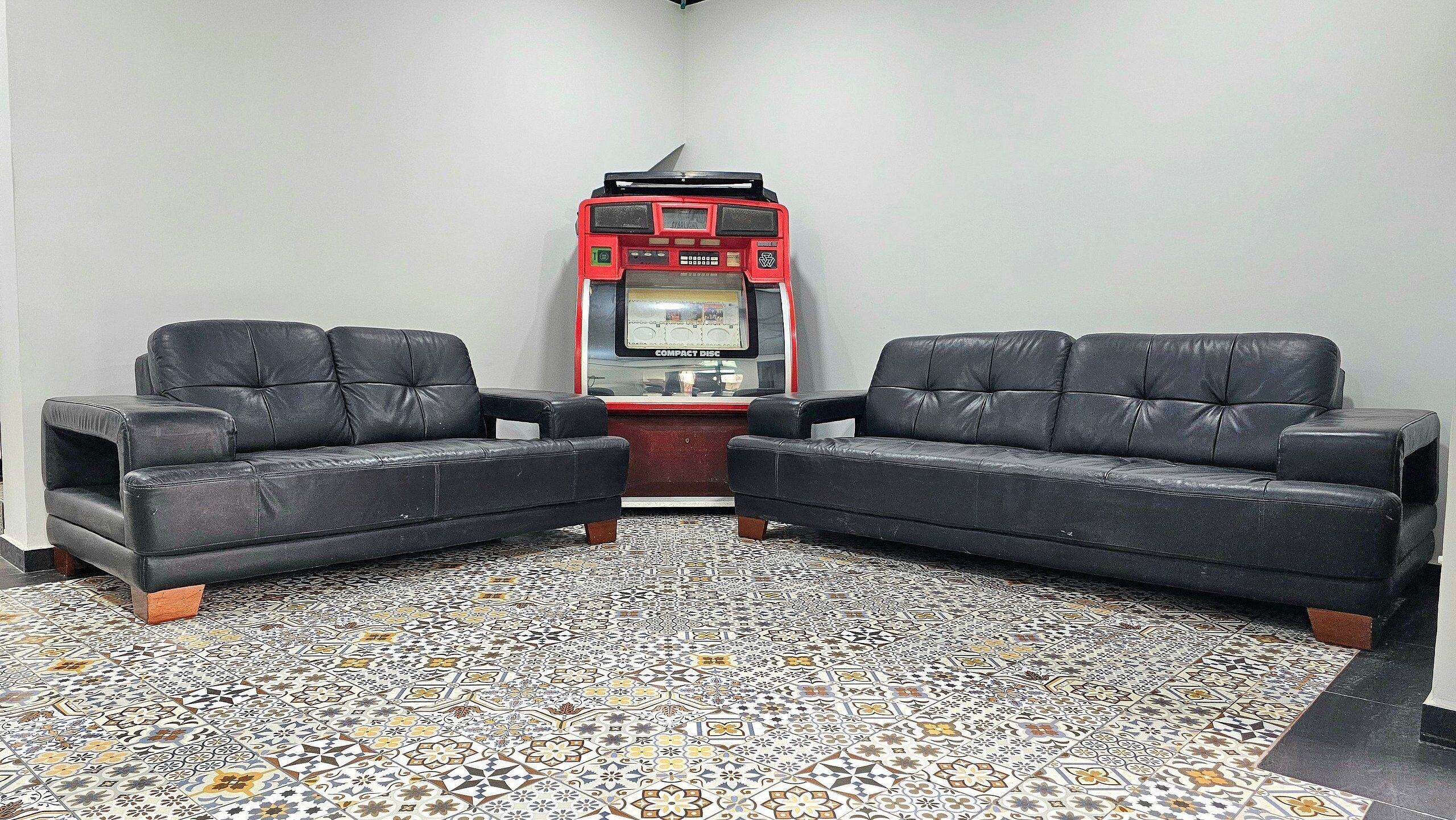 A room with two black leather sofas facing each other, a red and black arcade machine labeled "COMPACT DISC," and a patterned tile floor.