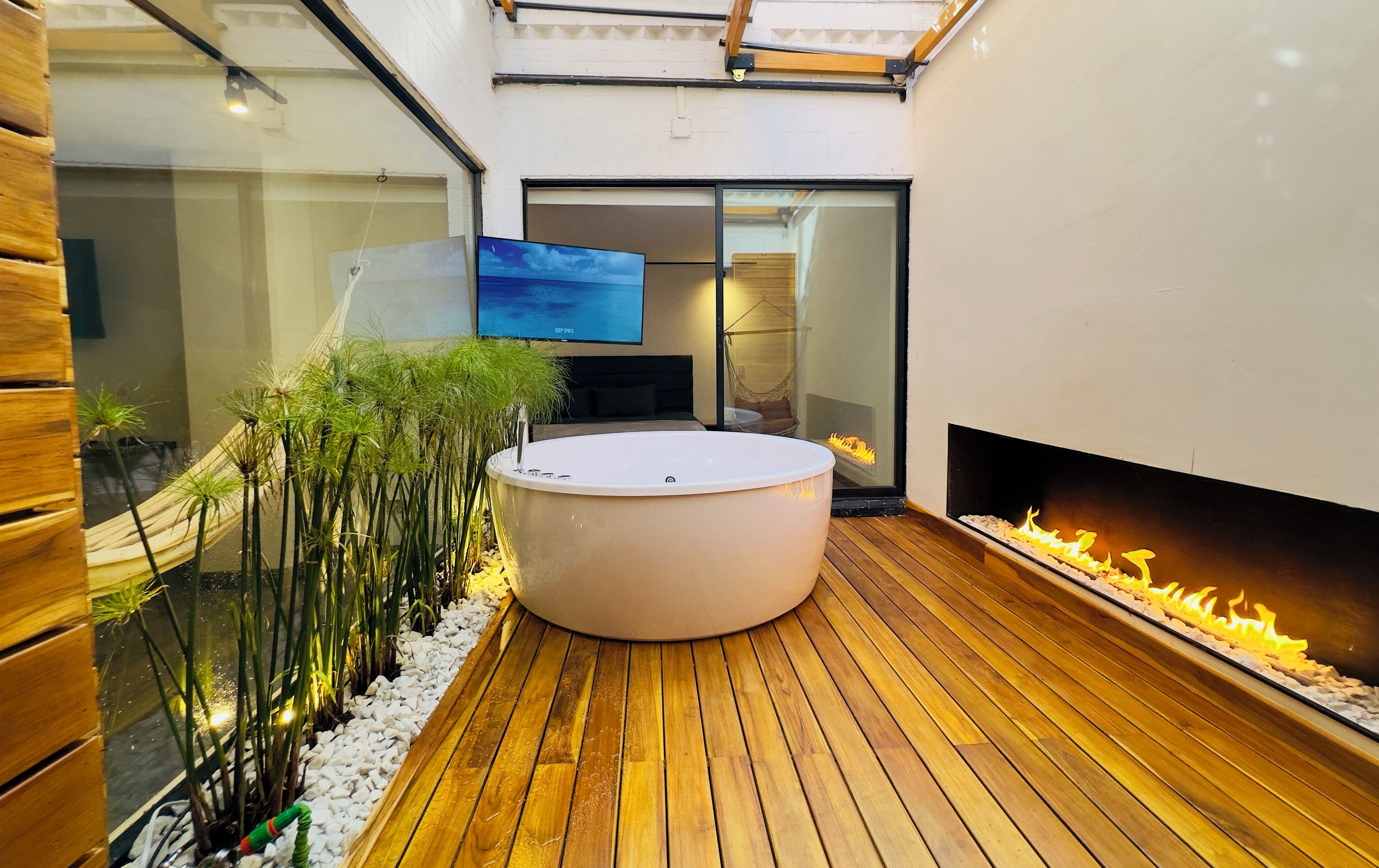 A modern outdoor living area with a wooden deck, a large round hot tub, a fire feature, and a television mounted on the wall.