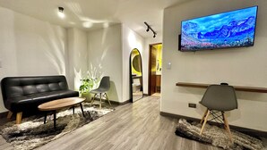 Un salon moderne avec un canapé en cuir noir, une table basse en bois et un fauteuil gris. Un téléviseur à écran plat est fixé au mur et affiche une image panoramique.