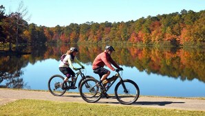 Dos ciclistas en un camino pavimentado junto a un lago tranquilo, con follaje otoñal al fondo.