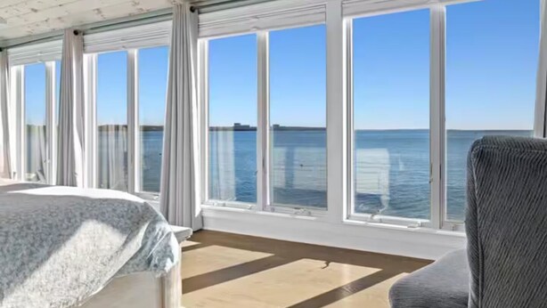 A room with a bed, a chair, and large windows overlooking the sea.