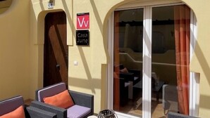 Un espacio de estar al aire libre techado con mobiliario de mimbre y cojines anaranjados, una puerta corredera de vidrio hacia un cuarto, y un letrero que dice 'Casa June'.