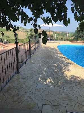 A stone-paved poolside area with a metal fence and a view of a hilly landscape in the background. - agriturismo cerquatonda (Montalto delle Marche)