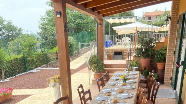 Une longue table à manger installée à l’extérieur, avec des chaises, une pergola au-dessus et des plantes en pot tout autour.