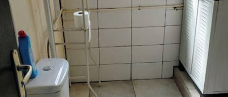Casa de banho pequena e compacta, com sanita, uma estante com produtos de higiene e uma janela com persianas.