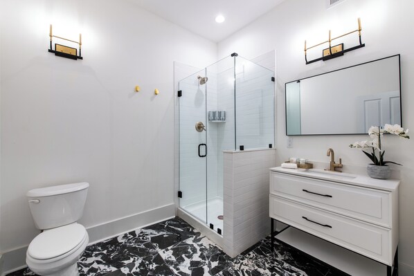Un baño moderno con ducha de vidrio, inodoro blanco, tocador blanco con cajones y un espejo grande.
