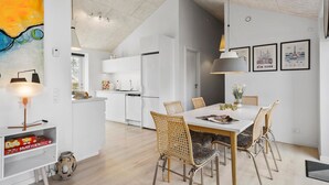 A modern kitchen with white cabinets, a dining area with a white table and wicker chairs, and a ceiling with exposed beams.