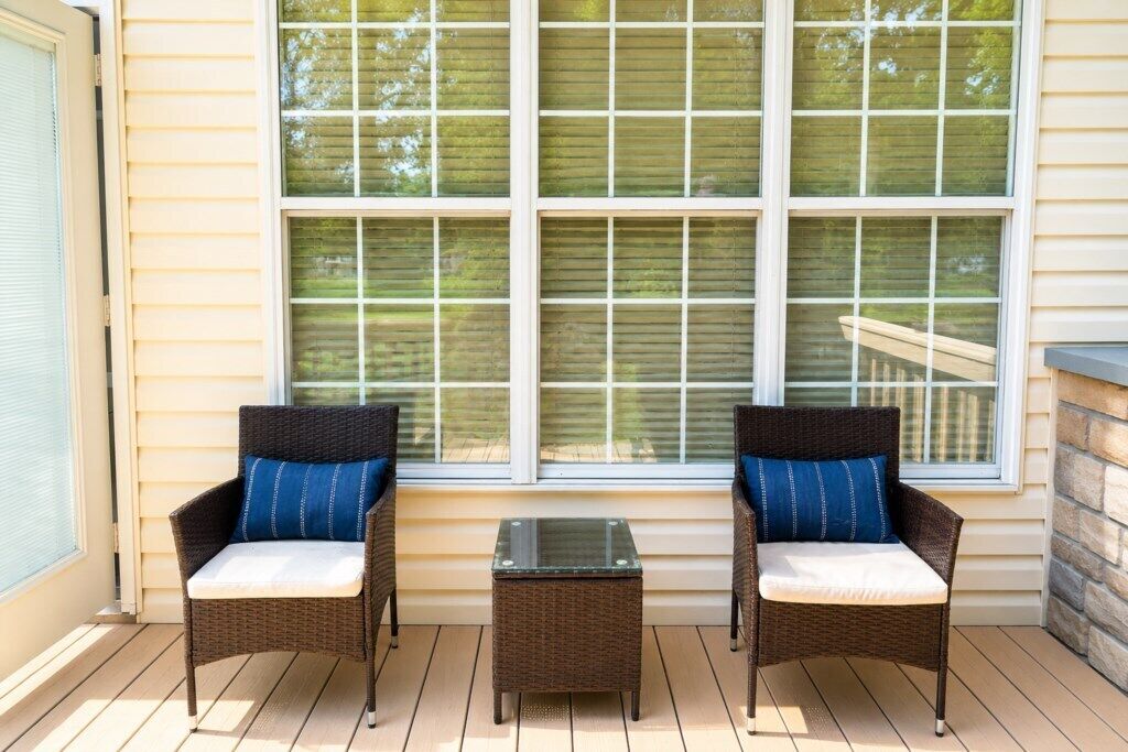 A covered porch with wicker chairs and a glass table, large windows, and a stone wall.