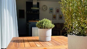 Et træbord med to plantede planter, en hvid gardin, og et køkken med et ur og billeder i baggrunden.