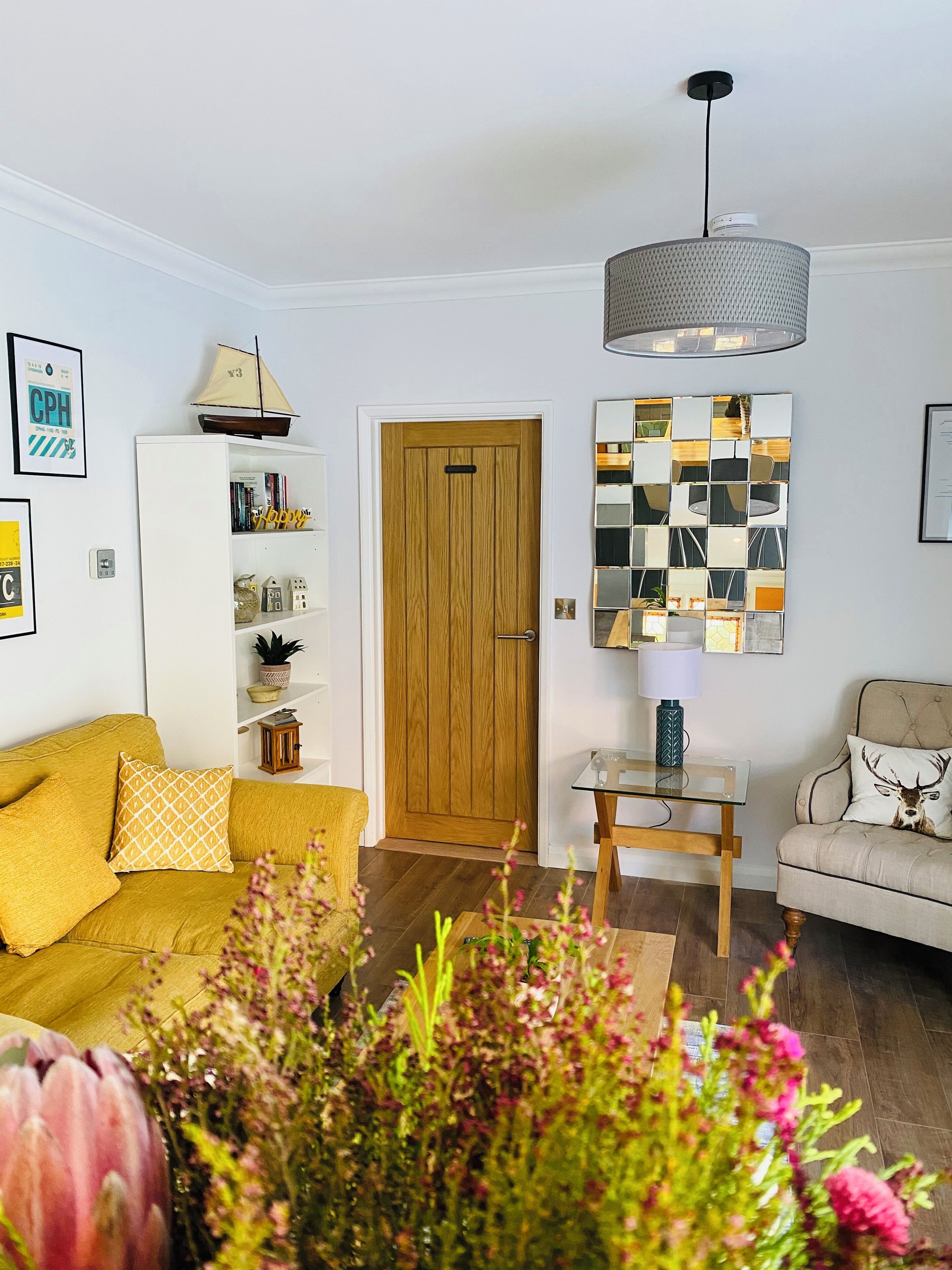A living room with a yellow sofa, a wooden door, a bookshelf, and a table with a lamp.