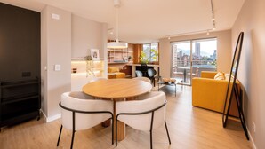 Sala de estar moderna com mesa de jantar de madeira, cadeiras brancas, sofá amarelo e uma grande janela com vista para a cidade.