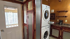 A laundry room with a washing machine and dryer, wooden cabinets, and a window with a view of trees.