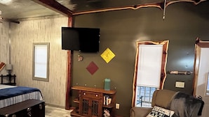 A living room with a flat-screen TV mounted on the wall, a wooden cabinet, a sofa with a cushion, and a patterned rug.