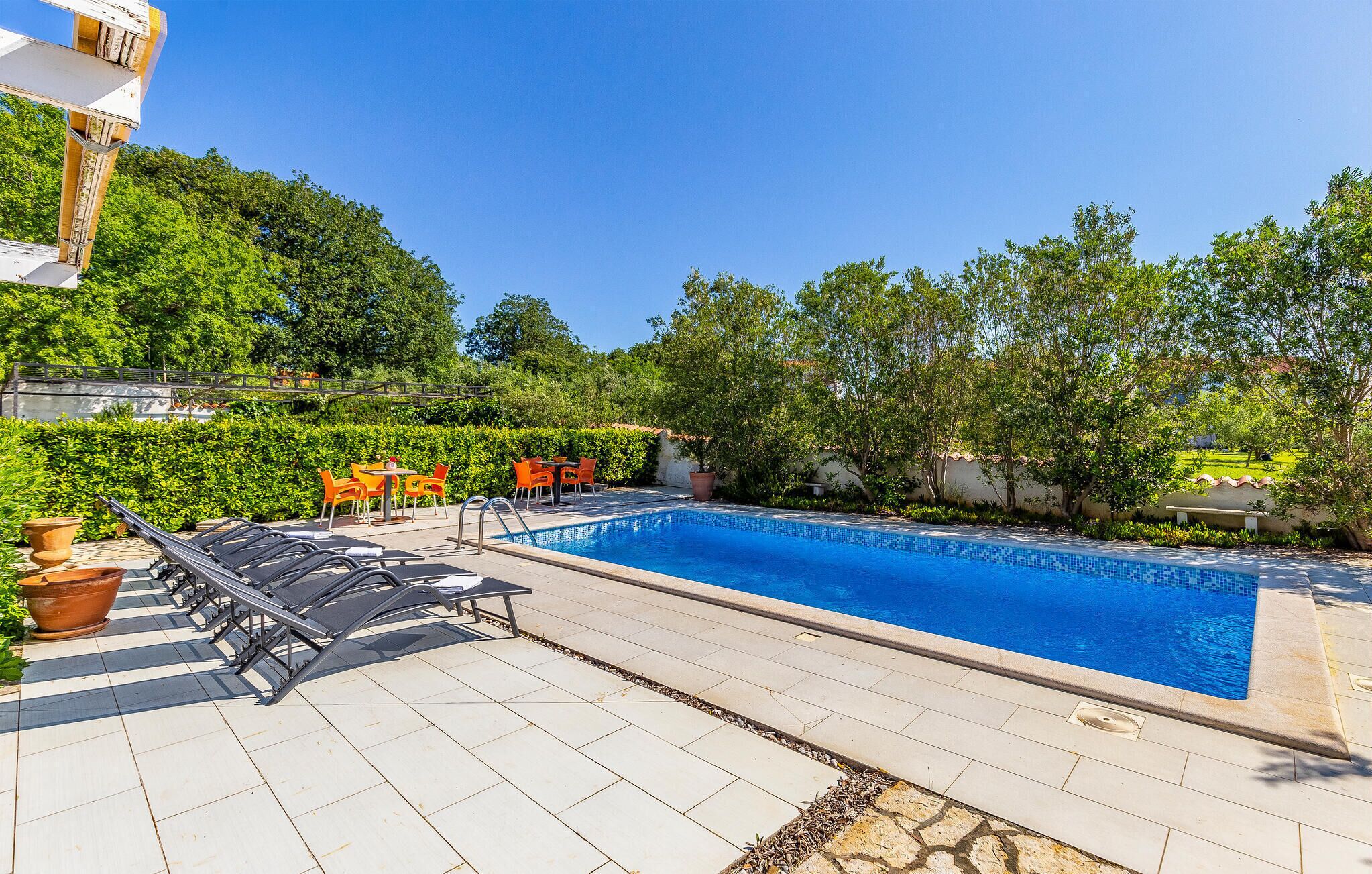A swimming pool with lounge chairs and tables, surrounded by greenery and a clear sky.