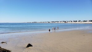 Una playa arenosa con algunas personas caminando, un mar tranquilo y casas al fondo.
