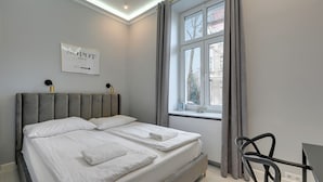 Une chambre moderne avec une tête de lit rembourrée de couleur grise, un lit avec du linge de lit blanc, une table de chevet et une fenêtre avec des rideaux.