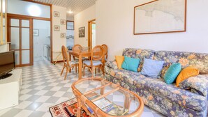 Un salon avec un sol à carreaux, un canapé à motifs floraux, une table basse en verre et un coin repas avec des chaises en osier.
