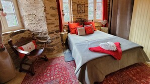 A rustic bedroom with a stone wall, a wooden bed with red and white pillows, a red patterned rug, and a window with curtains. - Le Manoir des Pélies-Au gré du vent. (Conques-en-Rouergue)
