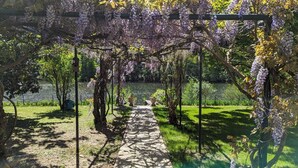 A garden pathway leading to a gazebo with purple flowers, surrounded by greenery and a body of water. - Le Manoir des Pélies-Au gré du vent. (Conques-en-Rouergue)