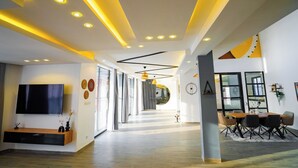 Eine moderne Hotelhalle mit Fernseher, einem hölzernen Empfangstresen, einem Essbereich mit Tisch und Stühlen sowie einem Gang zu den Zimmern.