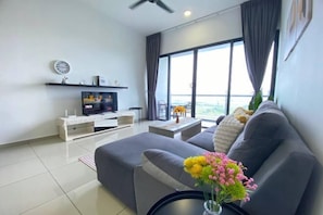 Ein modernes Wohnzimmer mit grauem Sofa, Flachbildfernseher, Couchtisch und großem Fenster mit Vorhängen.