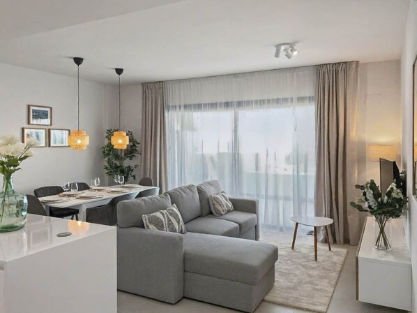 A modern living room with a grey sectional sofa, a dining area with a table set for two, and a large window with curtains.