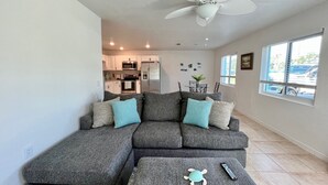 A modern living room with a grey sectional sofa, a white ceiling fan, a kitchen in the background, and a large window with a view of the outside.