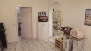 Un amplio salón con piso de baldosas, un acceso a otra habitación, un vitrina con adornos y una pequeña mesa con una casa modelo.