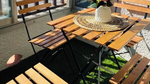 Ein Holztisch mit einer gewebten Tischmatte und einer Vase mit rosa Blumen auf einem Balkon mit Blick auf Gebäude.