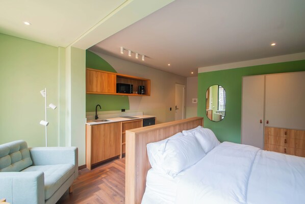 Una habitación de hotel con cama, una pequeña zona de estar, kitchenette y un baño visible a través de una puerta abierta.