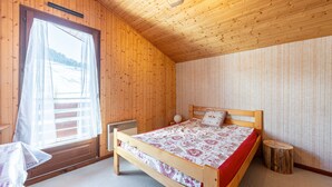 A wooden room with a bed, a window with curtains, a radiator, and a small stool.