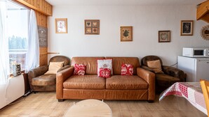 A cozy living room with a brown leather sofa, two armchairs, and a wooden coffee table. There are framed pictures on the wall, a white radiator, and a window with curtains.