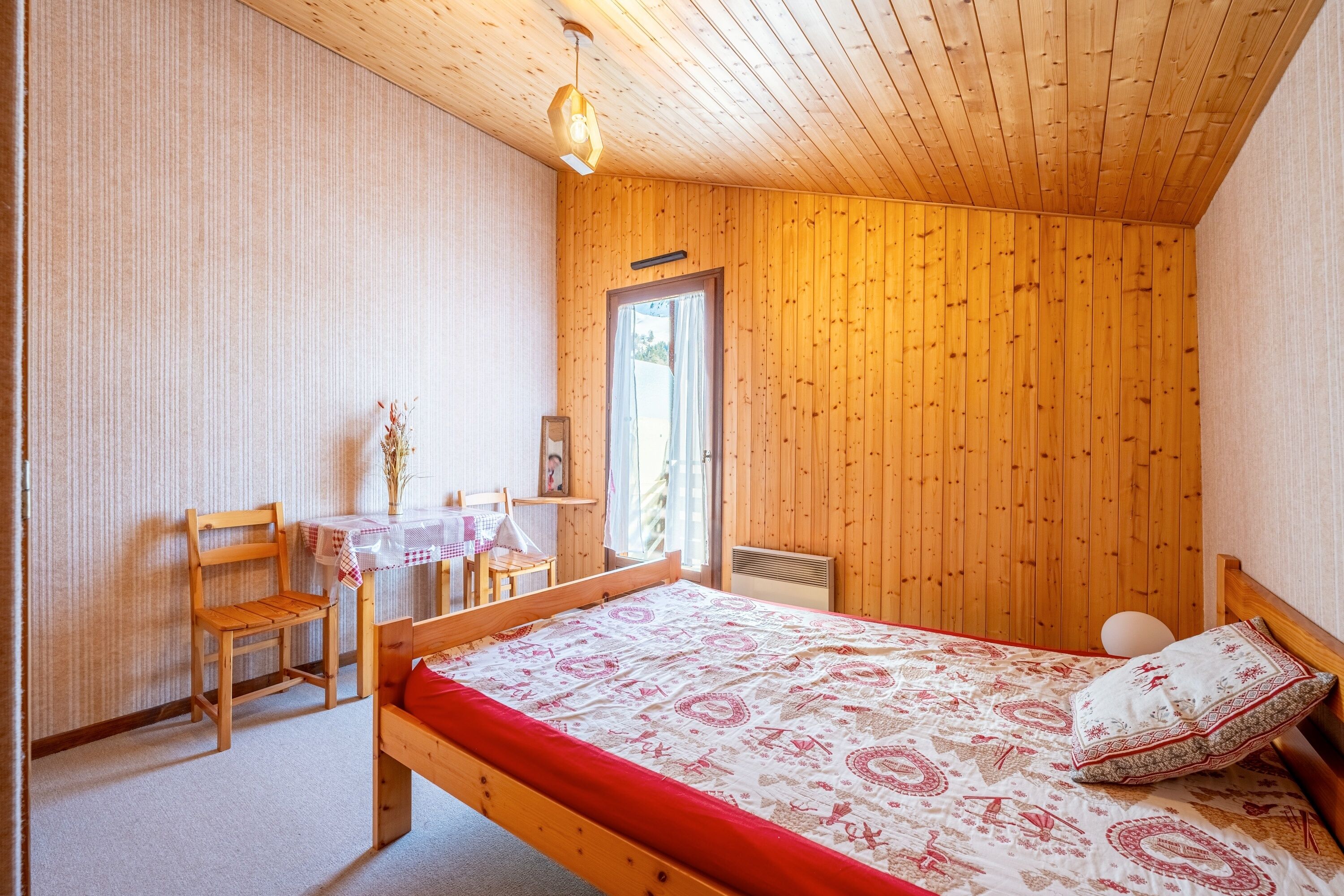 Une pièce aux murs lambrissés, contenant un lit, une chaise, une petite table et une fenêtre.