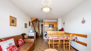 A cozy living room with a brown leather sofa, a dining table with chairs, and a kitchen area with wooden cabinets and a microwave.