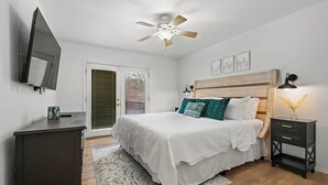 Un dormitorio con cama, ventilador de techo, mesita de noche, televisor montado en la pared y puerta que da a un balcón.