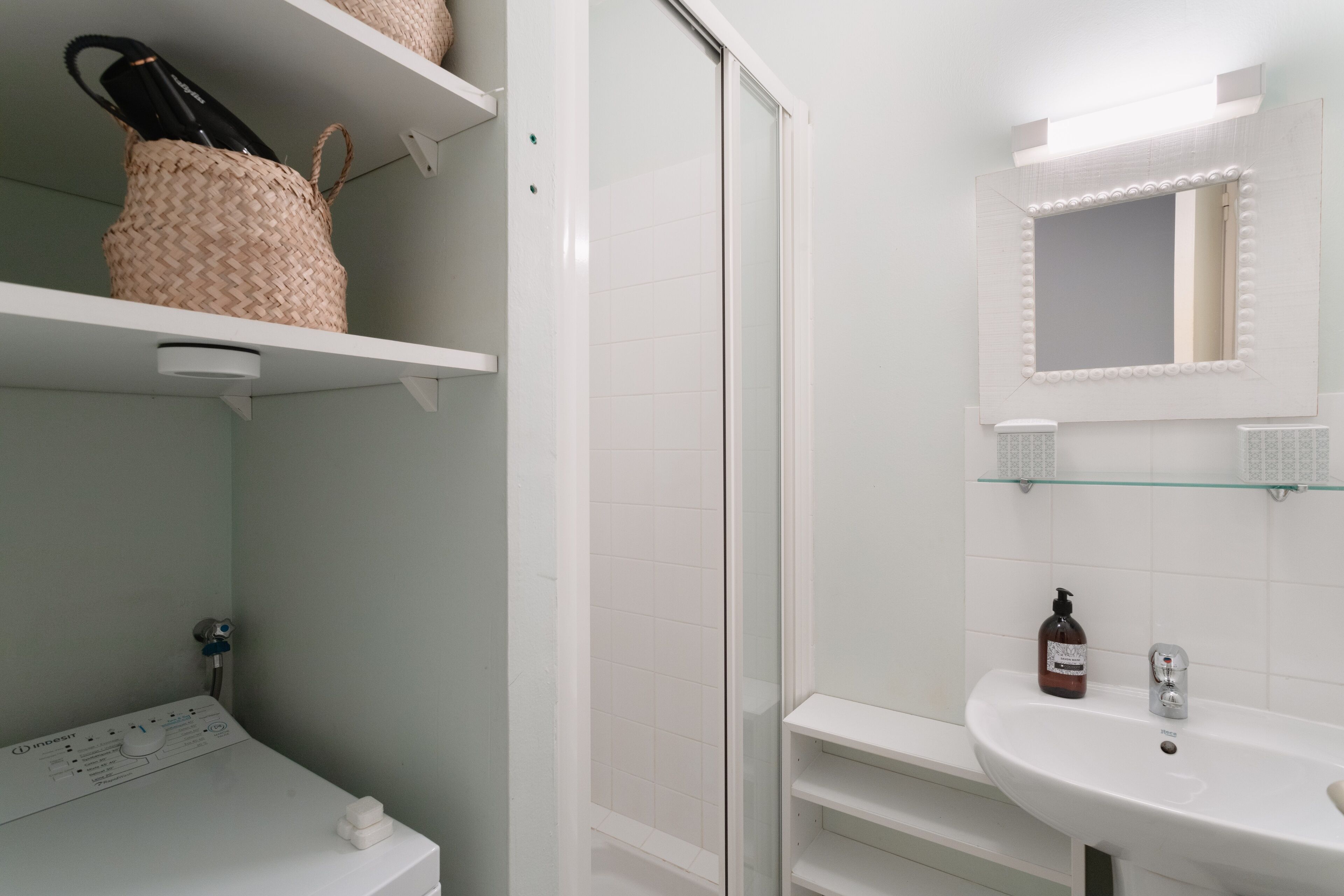 A bathroom with a white sink, a mirror with lights, a shower, and shelves with toiletries.