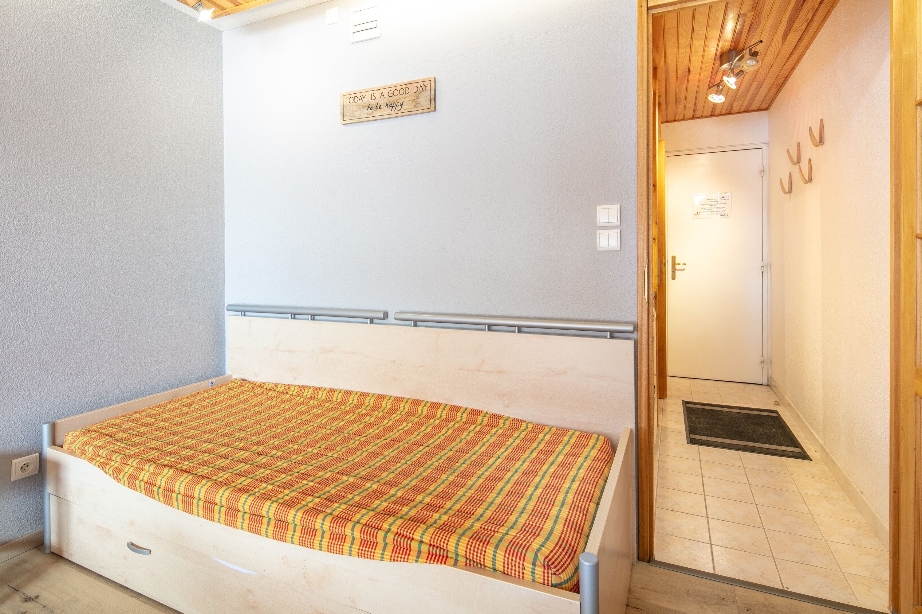 Un dormitorio con cama de madera, una mesita de noche pequeña, una lámpara de pared y una puerta con felpudo.