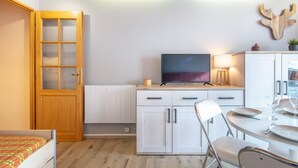 Une cuisine de taille réduite avec une porte en bois, un meuble blanc, une table à manger avec des chaises, un téléviseur et une plante fixée au mur.