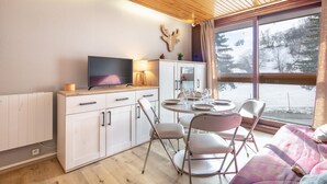 Une pièce chaleureuse avec un plafond en bois, une télévision, une table à manger avec des chaises et une fenêtre donnant sur des arbres et de la neige.