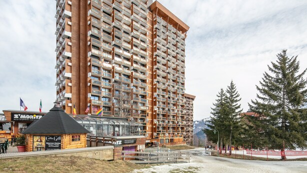 Un hôtel de grande hauteur doté de nombreux balcons, un restaurant appelé « Cotta Grill » situé à sa base, et une station de ski en arrière-plan.