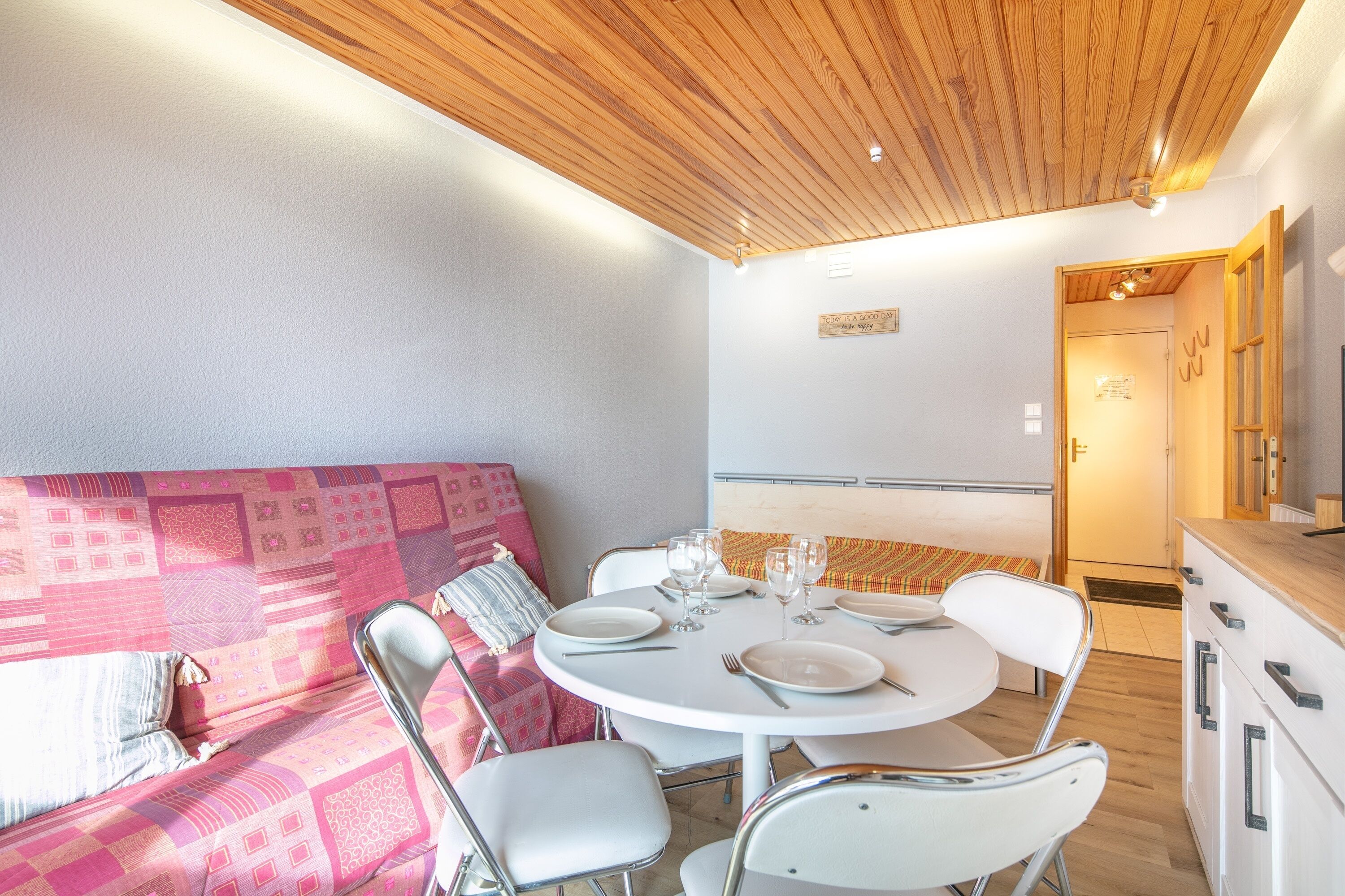 A cozy dining area with a round table set for four, surrounded by white chairs. A wooden ceiling with recessed lighting is visible above.