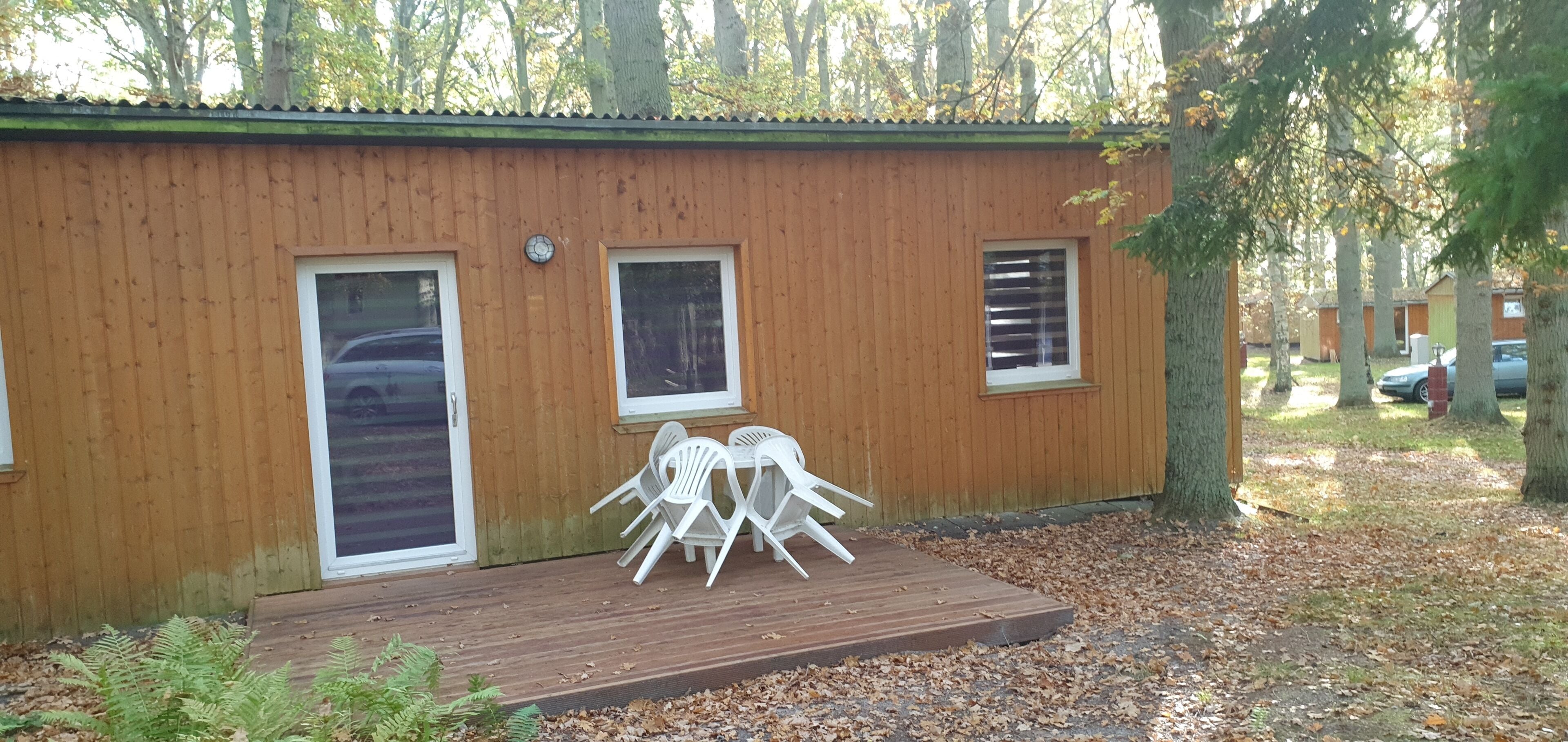 A wooden cabin with two windows and a door, a deck with white plastic chairs, and a forested background.