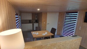 Habitación compacta con detalles de madera, una mesa de comedor con sillas, una zona de cocina y un ventanal grande con persianas.