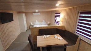 Una habitación pequeña con cama, una mesita con platos y tazas, un televisor montado en la pared y una ventana con cortinas a rayas.