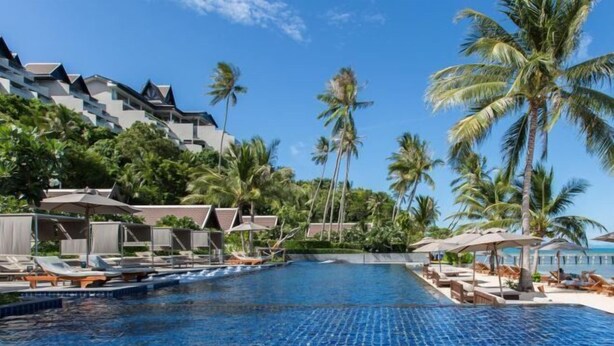 Ein Pool mit Liegestühlen und Sonnenschirmen, umgeben von Palmen und Hotelgebäuden.